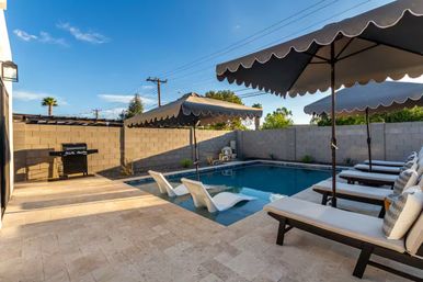Sunny private backyard pool patio with in‑pool loungers, cushioned chaise lounges under scalloped umbrellas, stone tile deck, gas grill and block privacy wall beneath a clear blue sky