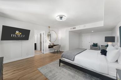 Bright contemporary bedroom suite with king bed in white linens and gray throw, wall-mounted TV, round gold mirror above a floating desk with upholstered chair, hardwood floors and a cozy seating area.