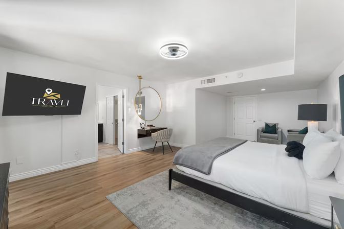 Bright contemporary bedroom suite with king bed in white linens and gray throw, wall-mounted TV, round gold mirror above a floating desk with upholstered chair, hardwood floors and a cozy seating area.