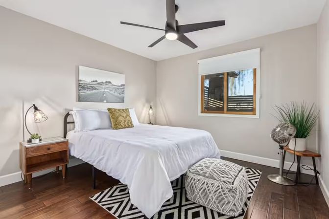 Modern bedroom with white bedding and yellow accent pillow, wooden nightstand and lamp, patterned rug and pouf, hardwood floors, ceiling fan, potted plant and window with natural light.