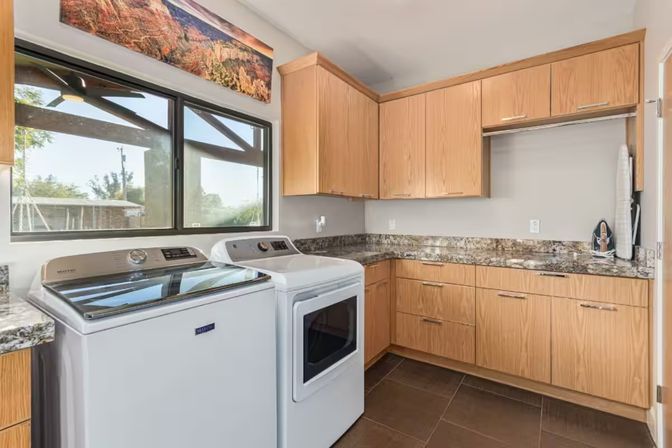 Bright modern laundry room with white washer and dryer, wood cabinets, granite countertops, large window overlooking backyard, tiled floor and ironing board.