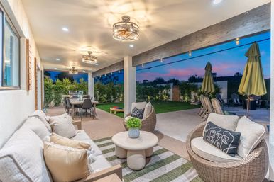 Cozy modern covered backyard patio at sunset with wicker lounge chairs, cushioned sofa, round coffee table, dining table, string lights and landscaped lawn.