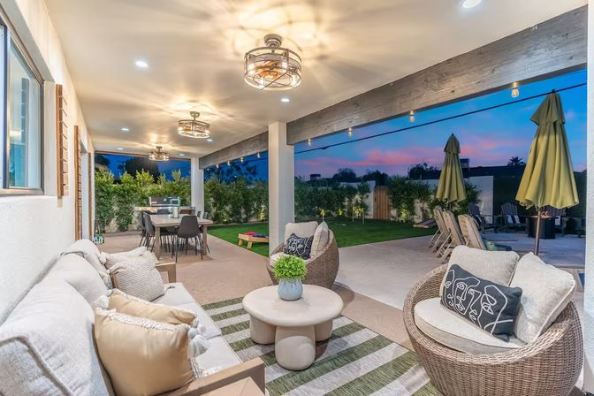 Cozy modern covered backyard patio at sunset with wicker lounge chairs, cushioned sofa, round coffee table, dining table, string lights and landscaped lawn.
