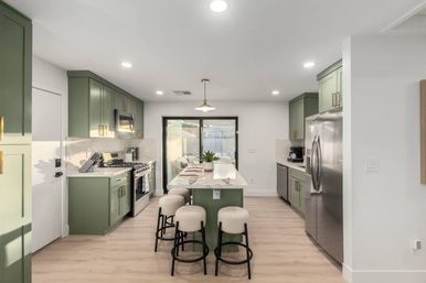 Bright modern open-concept kitchen with sage green cabinets, quartz island with four round upholstered stools, stainless steel appliances, sliding glass patio doors to backyard, and light wood floors.