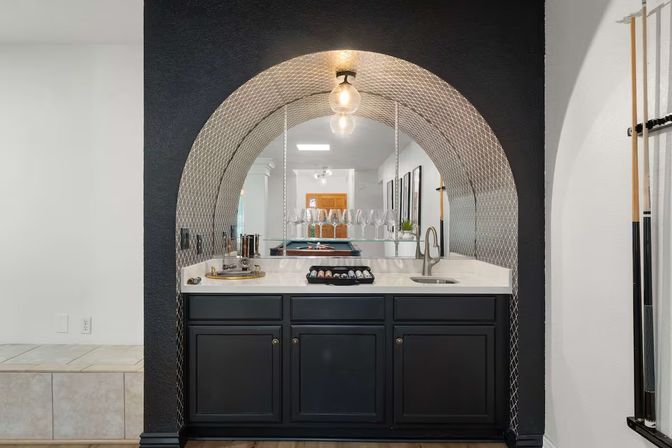 Modern home wet bar alcove with decorative arched mirrored backsplash and exposed bulb pendant, white quartz countertop with sink and glassware, dark cabinetry, and a reflected pool table in a residential game room.