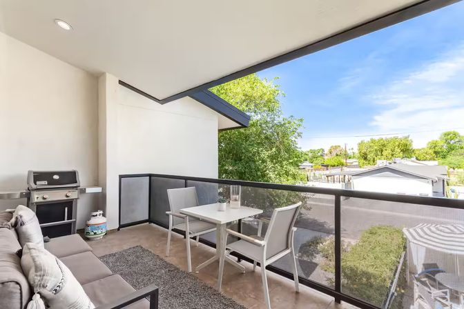 Covered modern balcony with cushioned sofa, small white dining table and two chairs, propane grill and rug, glass railing overlooking suburban rooftops and green trees under a bright blue sky.
