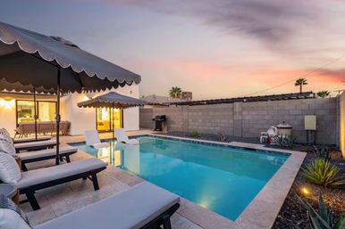 Backyard swimming pool at sunset with turquoise water, cushioned lounge chairs under scalloped umbrellas, two in-pool loungers, modern white home, block wall fence, desert landscaping with agave plants and a barbecue.
