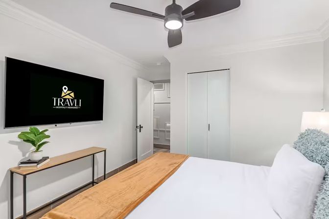 Bright modern bedroom interior with wall-mounted flat-screen TV, ceiling fan, crisp white linens with tan throw, slim console table with potted plant and sliding closet doors.