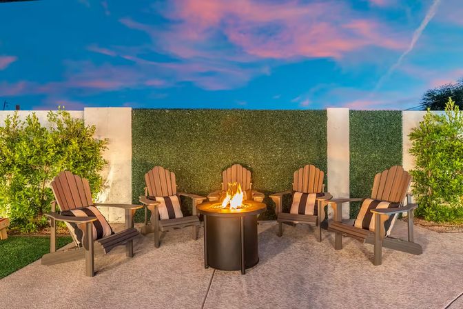 Cozy backyard patio at twilight with five Adirondack chairs and striped cushions around a glowing outdoor fire pit, green hedge wall and pink-blue sky.