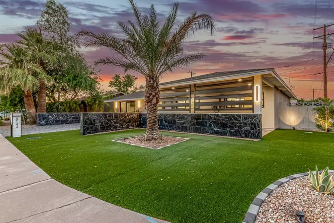 Sunset exterior of a single-story mid-century modern home with a central palm tree, black stone accent wall, neat green lawn (artificial turf) and agave in a desert-style front yard.