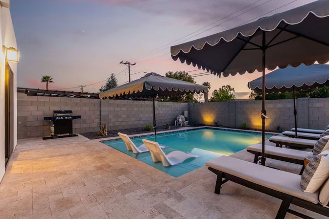 Backyard pool at sunset with two in-pool loungers, cushioned chaise lounges under scalloped umbrellas, tiled patio, gas grill and warm wall accent lighting.