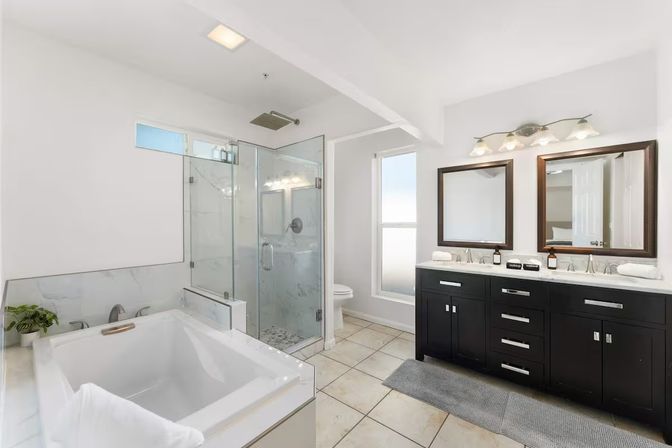 Spa-like modern bathroom with a white soaking tub, frameless glass rainfall shower, double-sink dark wood vanity with twin mirrors, marble accents, frosted window and large beige floor tiles.