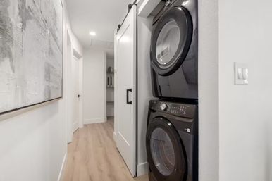 Modern hallway with light wood floors and white walls, showing a sliding-barn-door laundry closet with a sleek stacked black front-loading washer and dryer and abstract wall art.