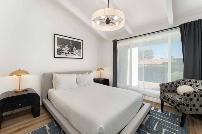 Bright modern bedroom with low platform bed and crisp white linens, gold bedside lamps, patterned accent chair, pendant light and sliding glass door to a sunny suburban yard