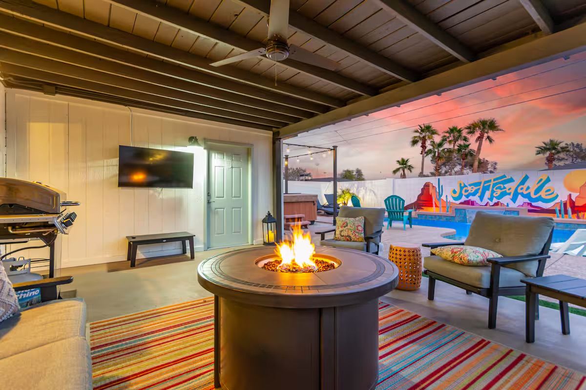 Cozy covered backyard patio at sunset with a round gas fire pit, cushioned lounge chairs, striped rug, wall-mounted TV and grill, overlooking a pool, hot tub and colorful mural with palm trees.