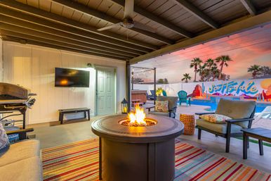 Cozy covered backyard patio at sunset with a round gas fire pit, cushioned lounge chairs, striped rug, wall-mounted TV and grill, overlooking a pool, hot tub and colorful mural with palm trees.