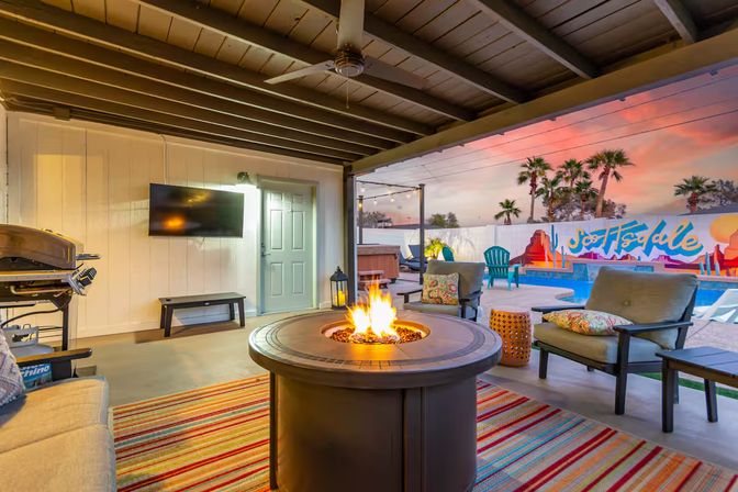Cozy covered backyard patio at sunset with a round gas fire pit, cushioned lounge chairs, striped rug, wall-mounted TV and grill, overlooking a pool, hot tub and colorful mural with palm trees.
