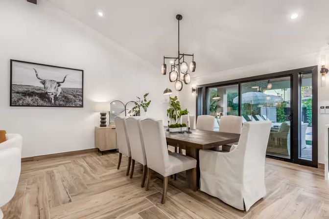Sunlit modern dining room with a rustic wooden table, eight beige slipcovered chairs, globe pendant chandelier, light wood plank floors and sliding glass doors opening to a poolside patio with umbrella and outdoor seating, framed highland cow art on the wall.