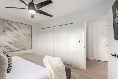 Bright, airy modern bedroom with a sleek black ceiling fan, large abstract wall art, white double-closet doors, light wood floors and a cozy bed layered with neutral linens and a textured throw.