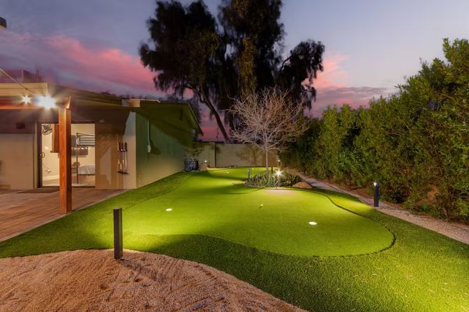 Twilight residential backyard putting green with artificial turf, built-in golf cups, pathway lights and a covered patio next to a tall privacy hedge and decorative tree.