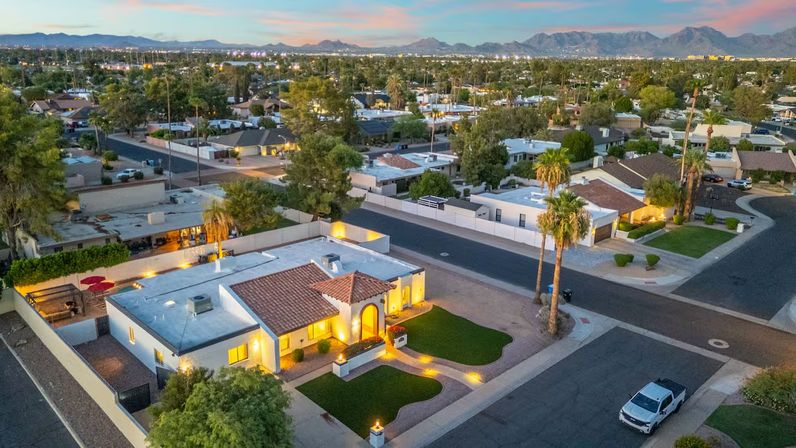 Aerial dusk view of a glowing Spanish-style single-story home with red tile roof, lit pathways, palm trees and manicured lawns in a suburban desert neighborhood with mountains on the horizon.