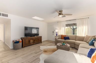 Bright sunlit living room with a large beige sectional sofa, colorful throw pillows, round woven coffee table, wooden media console with flat-screen TV, ceiling fan, light wood floors and sheer curtains — cozy and modern.