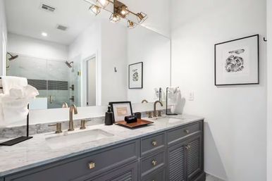 Bright contemporary master bathroom with marble double vanity, gray cabinets, brass faucets, large mirror, modern light fixture and glass-enclosed shower