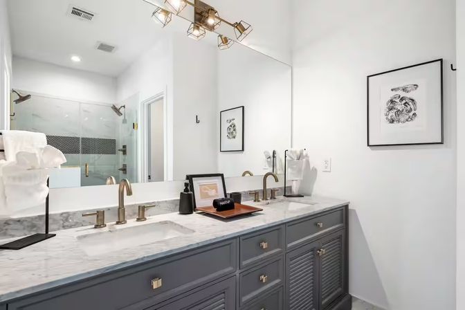 Bright contemporary master bathroom with marble double vanity, gray cabinets, brass faucets, large mirror, modern light fixture and glass-enclosed shower