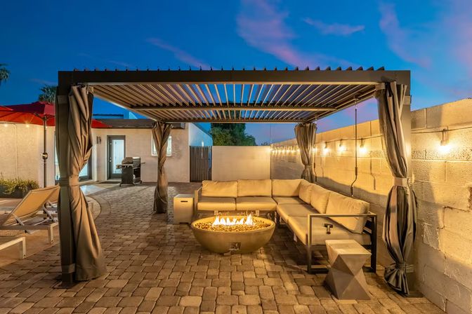 Dusk backyard patio with louvered pergola, cozy beige sectional sofa, round gas fire pit on paver flooring, string lights and curtained columns creating an inviting outdoor lounge.