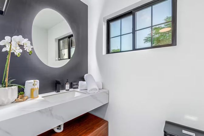 Modern minimalist bathroom vanity with white marble countertop, round mirror on a black accent wall, black faucet, orchid plant, folded towels and a grid window letting in natural light.