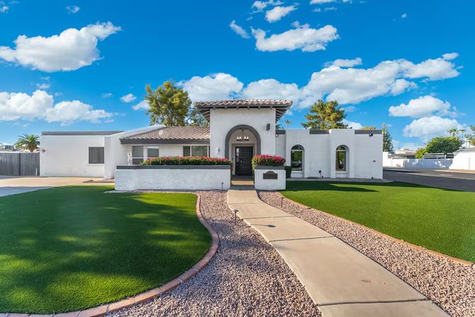White Southwestern-style single-story stucco home with Spanish tile roof and arched entry, colorful flower beds, curved concrete walkway bordered by gravel and lush green turf lawns, set under a bright blue sky with puffy clouds.