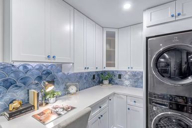 Modern residential laundry room with white shaker cabinets, blue scalloped fish-scale tile backsplash, built-in white countertop workspace and stacked stainless steel washer and dryer, styled with a desk lamp, books, clock and potted plant.