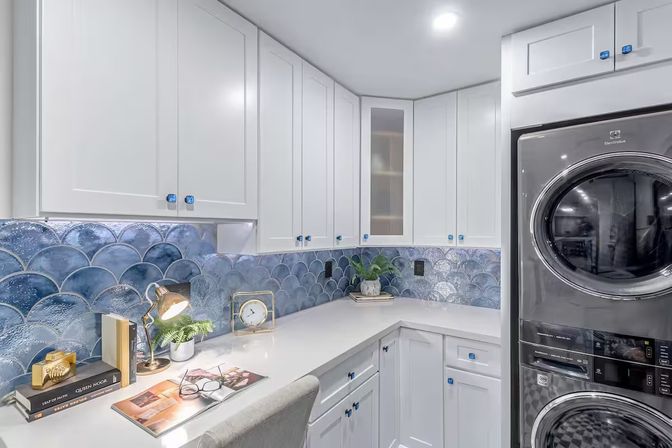 Modern residential laundry room with white shaker cabinets, blue scalloped fish-scale tile backsplash, built-in white countertop workspace and stacked stainless steel washer and dryer, styled with a desk lamp, books, clock and potted plant.