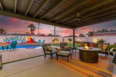 Cozy covered backyard patio at sunset — cushioned lounge chairs around a lit round fire pit on a striped rug, swimming pool with turquoise Adirondack chairs, colorful desert mural on the wall and palm trees.