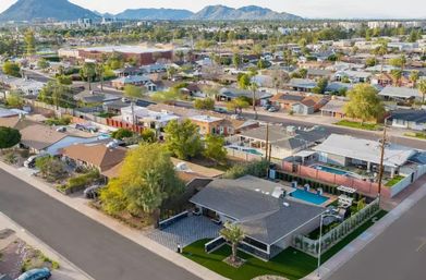 Aerial view of a sunny suburban neighborhood with single-story ranch homes, backyard pools, palm trees and mountain skyline in the distance