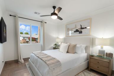 Bright, airy modern bedroom with a queen bed in neutral linens, textured throw and accent pillows, wood nightstands with lamps, abstract wall art, black ceiling fan, hardwood floors and a curtained window overlooking a suburban street.