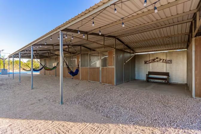 Covered stable-style shelter with metal roof and stall panels, colorful hammocks hung between posts, string lights overhead, gravel floor and a bench against a metal wall in a sunny desert ranch setting.