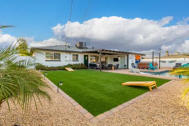 Sunny Arizona backyard with green artificial turf and cornhole boards, gravel desert landscaping, covered patio dining area, pool and hot tub with turquoise lounge chairs, palm plants and blue sky clouds.