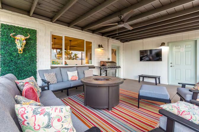 Covered backyard patio lounge with gray sectional sofas and colorful patterned pillows, striped rug, round fire‑pit coffee table, wall‑mounted TV, grill, ceiling fan, and faux greenery wall with decorative bull skull