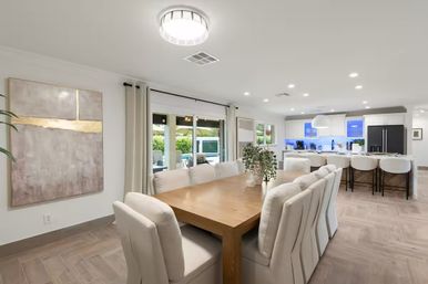 Bright modern open-plan dining room and kitchen featuring a long wooden table with beige upholstered chairs, a simple greenery centerpiece, sliding glass doors to a pool-side patio, and a white kitchen island with bar stools and stainless appliances.