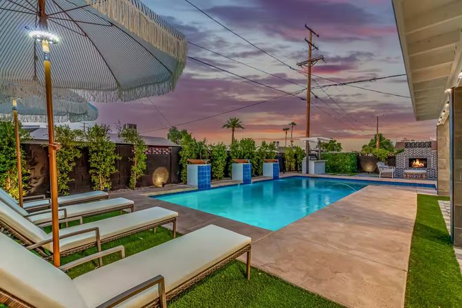Residential backyard pool at sunset with blue-tiled water features, cushioned lounge chairs under fringed umbrellas, palm trees and an outdoor fireplace.