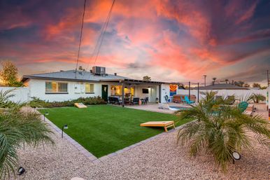 Suburban Arizona backyard at sunset with vibrant pink-orange sky, artificial turf lawn edged by gravel and palm plants, cornhole boards on the grass, small pool and hot tub, and a covered patio dining area with an Arizona flag mural.
