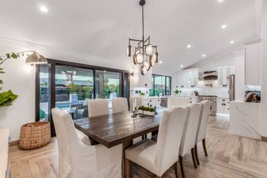 Sunlit modern open-plan dining room and kitchen with a rustic dark wood table, cream upholstered chairs, pendant chandelier, sliding glass doors to a backyard pool, stainless steel appliances, marble countertops, and wide-plank wood floors.