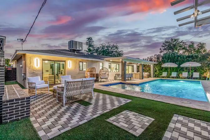 Inviting modern suburban backyard at dusk with a lit rectangular swimming pool, poolside loungers and umbrellas, wicker lounge seating on patterned pavers, covered patio dining and hanging egg chairs under a purple-pink sunset sky.
