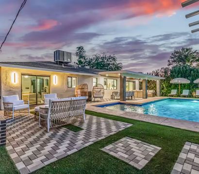Inviting modern suburban backyard at dusk with a lit rectangular swimming pool, poolside loungers and umbrellas, wicker lounge seating on patterned pavers, covered patio dining and hanging egg chairs under a purple-pink sunset sky.