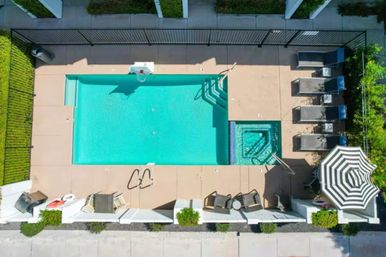 Aerial view of a turquoise rectangular swimming pool and adjacent square hot tub in a residential courtyard, with pool deck, lounge chairs, striped umbrella, fencing and surrounding greenery.