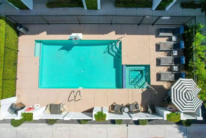 Aerial view of a turquoise rectangular swimming pool and adjacent square hot tub in a residential courtyard, with pool deck, lounge chairs, striped umbrella, fencing and surrounding greenery.