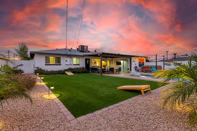 Arizona backyard at vibrant sunset with artificial turf lawn and cornhole boards, pebble landscaping, pool and hot tub, covered patio dining area with string lights and lounge seating.