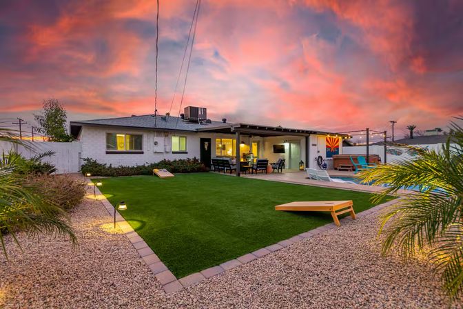 Arizona backyard at vibrant sunset with artificial turf lawn and cornhole boards, pebble landscaping, pool and hot tub, covered patio dining area with string lights and lounge seating.