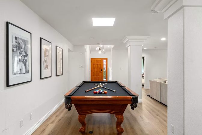Modern home game room with a wooden pool table topped with black felt on light hardwood floors, white walls with framed black-and-white portraits, an orange wooden entry door in the background and an open living area with a beige sofa.